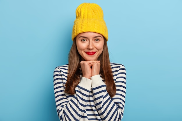 Tender good looking dark haired woman stands against blue background in yellow hat and striped sweater, wears red lipstick