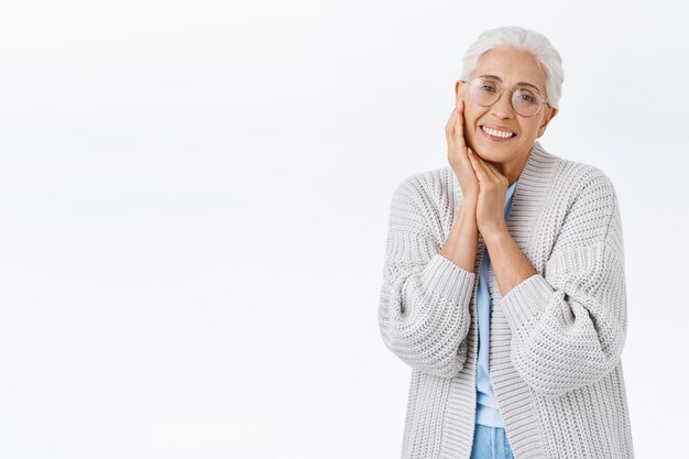 Tender lovely senior woman with grey combed hair in prescription glasses, cozy cardigan, touching cheek and smiling heartwarming