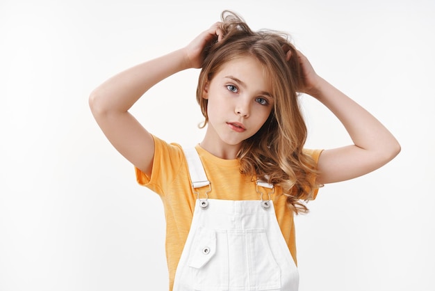 Tender lovely young blond girl in summer dungarees over yellow t-shirt, look thoughtful and cute camera, scratch hair bothered, touch hairstyle, suffer head lice