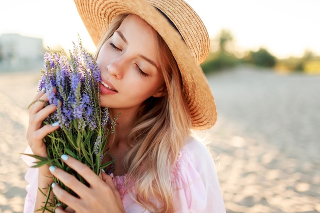 Tender pretty woman  in  straw hat posing on sunny beach near ocean with bouquet of flowers. Close up portrait.