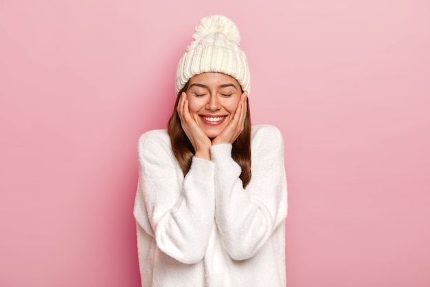 Tender relaxed carefree woman with toothy smile, wears white casual jumper and hat, feels joyful and unbothered, keeps eyes closed, poses against pink background