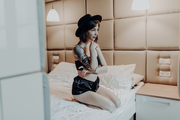 Tender tiny woman in fashion black underwear and wide hat sit on the bed in the morning