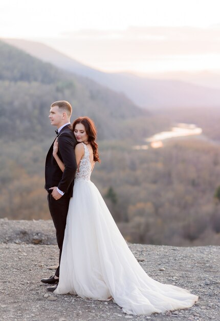 Tender wedding couple on the dusk on the top of a hill is hugging, dressed in luxury wedding attire