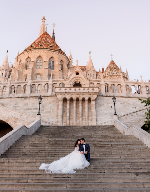 Tender wedding couple is sitting on the stairs of an old stone historical building on the warm evening