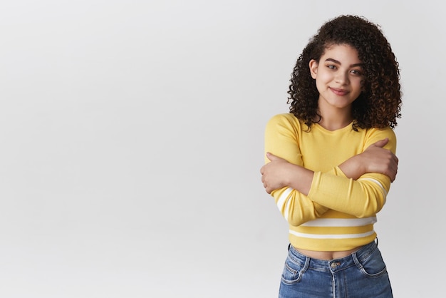 Tenderness wellbeing beauty concept Attractive lovely curlyhaired young woman hugging herself wearing cropped sweater embracing body smiling cute gentle dream romantically white background