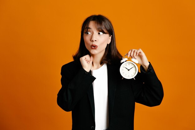 Tense looking at side holding alarm clock young beautiful female wearing black jacket isolated on orange background