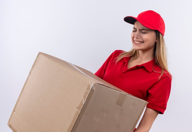 Tense young delivery girl wearing red uniform and cap holding heavy box isolated on white