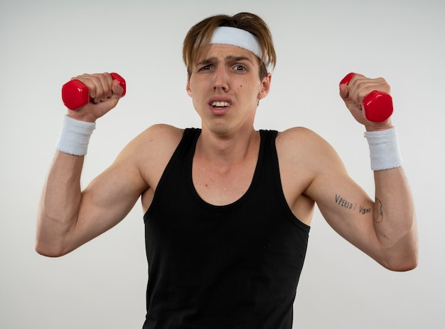 Tense young sporty guy wearing headband and wristband exercising with dumbbells isolated on white wall