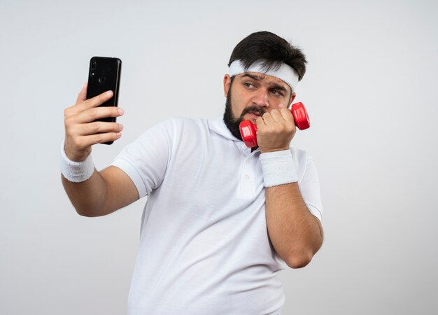 Tense young sporty man wearing headband and wristband exercising with dumbbell take a selfie isolated on white wall