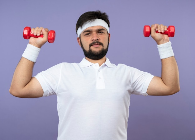 Tense young sporty man wearing headband and wristband exercising with dumbbells