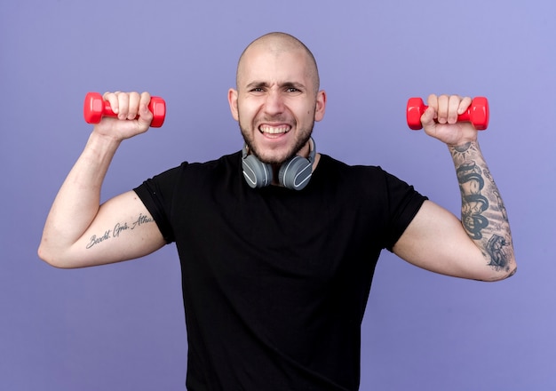 Free photo tense young sporty man wearing headphones on neck exercising with dumbbells isolated on purple