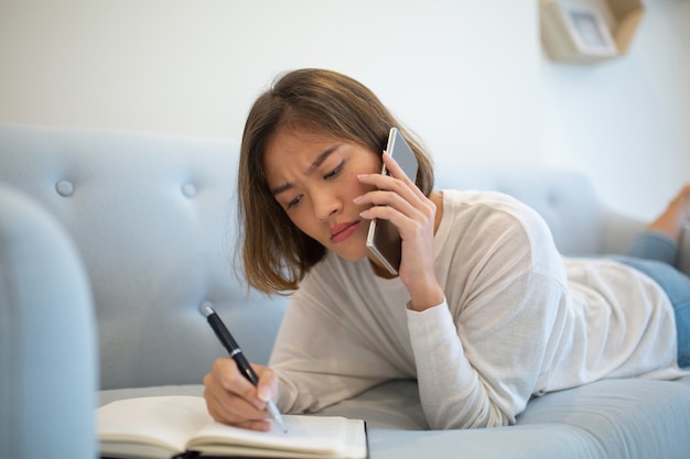 Tensed lady making notes and calling on phone on sofa