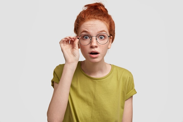 Free photo terrific ginger woman with stunned expression, stares through eyewear, opens mouth slightly, sees something surprising