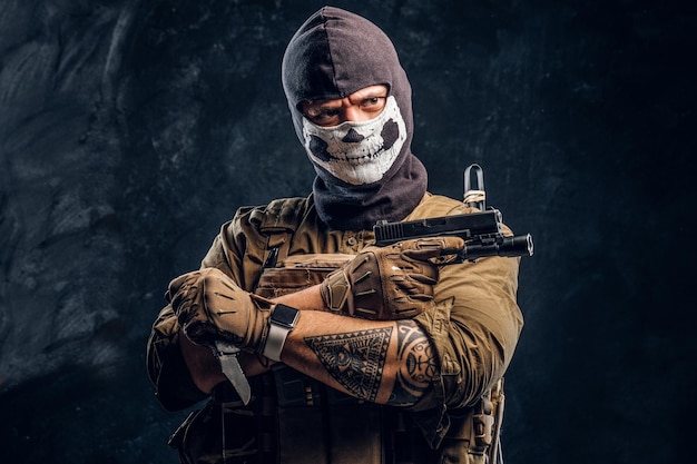 Free Photo a terrorist in a military uniform and a skull balaclava holding a pistol and a knife and looks at the camera with a menacing look. studio photo against a dark textured wall