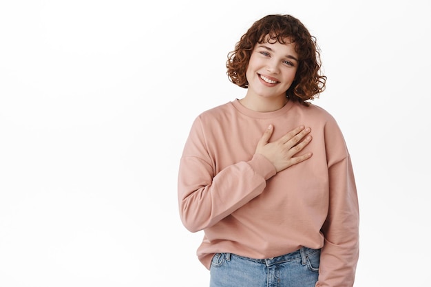 Thankful friendly girl, hold hand on heart and smile touched, appreciate nice gesture, thanking for something, being grateful, standing against white background.