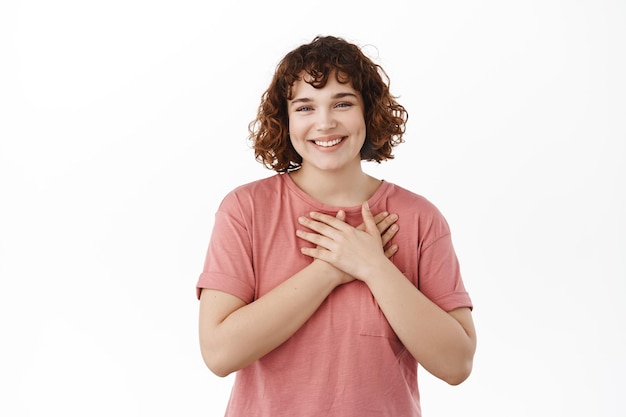 Free photo thankful girl appreciate nice gesture, holding hands on heart, smiling grateful, express gratitude, being heartfelt and touched, standing in t-shirt over white background