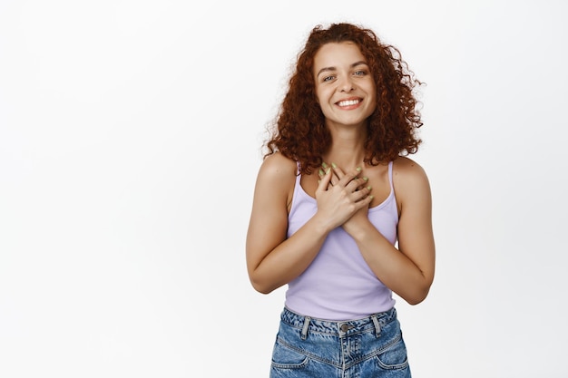 Free photo thankful smiling redhead woman with curly hair, holding hands near chest and looking grateful, flattered with compliment, pleased of something good, white background.