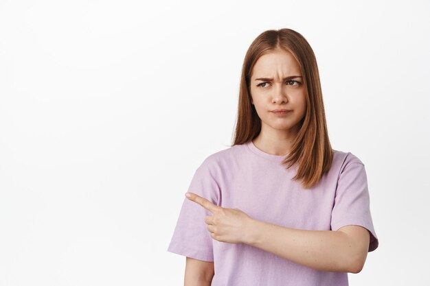 That it bad. Displeased blond woman pointing, furrow eyebrows with dislike, staring at smth with judging face, showing awful banner logo on left side, complaining, white background