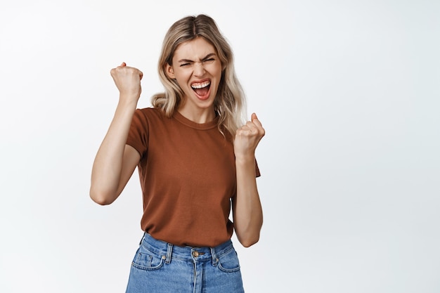 Thats the spirit. Enthusiastic blond girl winning, celebrating victory, scream from excitement and triumphing, standing on white