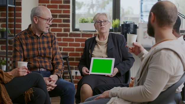 Therapist holding tablet with horizontal green screen at aa meeting with people. Woman psychiatrist showing chroma key and isolated mockup template on display to patients with addiction.