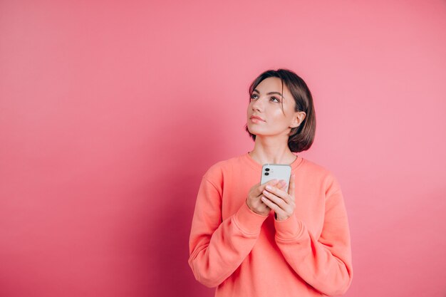 Thinking dreaming young beautiful woman posing isolated over pink wall background using mobile phone