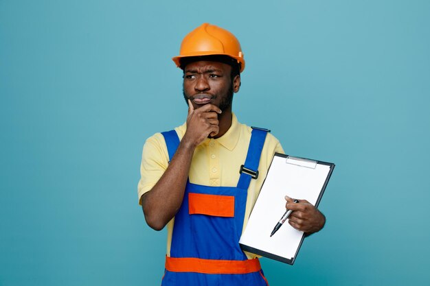 Thinking grabbed chin young african american builder in uniform holding clipboard isolated on blue background