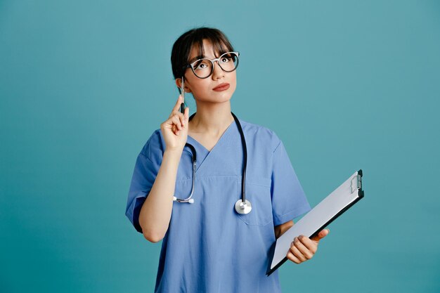 Thinking holding clipboard with pen young female doctor wearing uniform fith stethoscope isolated on blue background