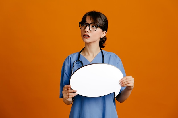 Thinking holding speech bubble young female doctor wearing uniform fith stethoscope isolated on orange background