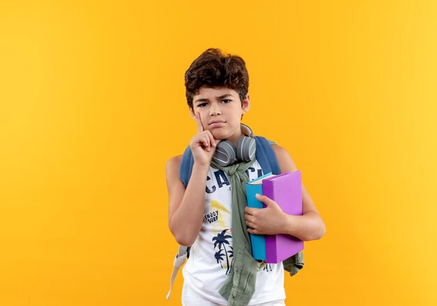 Thinking little school boy wearing back bag and headphones holding books and putting hand on cheek isolated on yellow