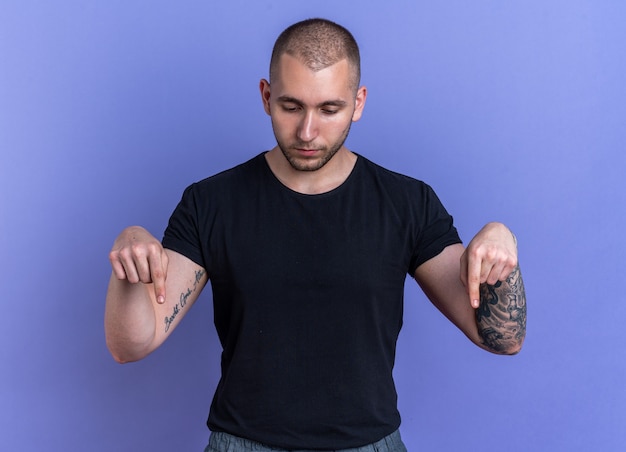 Free photo thinking looking down young handsome guy wearing black t-shirt points at down isolated on blue wall