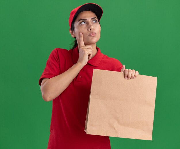 Thinking looking at side young delivery girl wearing uniform and cap holding paper food package putting finger on cheek isolated on green wall