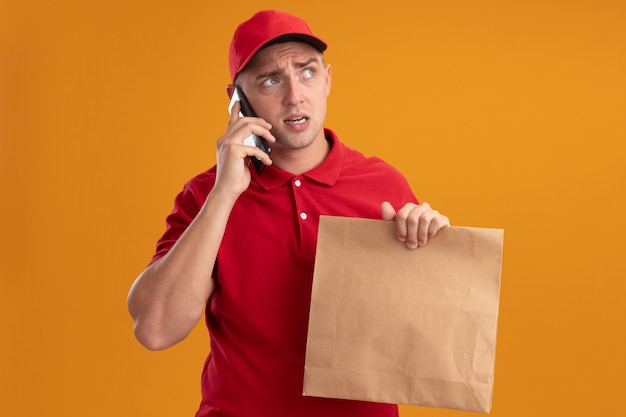 Thinking looking at side young delivery man wearing uniform with cap holding paper food package speaks on phone isolated on orange wall