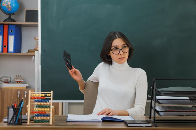 thinking looking at side young female teacher wearing glasses holding calculator sitting at desk with school tools on in classroom