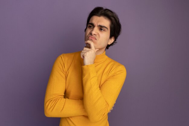 Thinking looking at side young handsome guy wearing yellow turtleneck sweater putting hand on chin isolated on purple wall