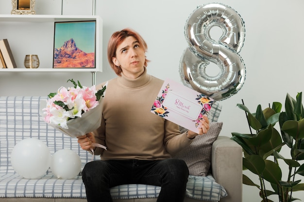 Thinking looking up handsome guy on happy women day holding bouquet with greeting card sitting on sofa in living room