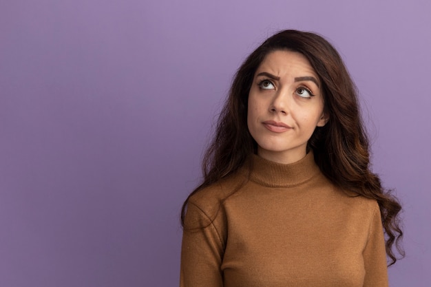 Free photo thinking looking up young beautiful girl wearing brown turtleneck sweater isolated on purple wall with copy space