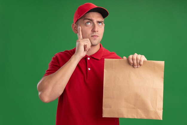 Thinking looking up young delivery man wearing uniform and cap holding paper food package putting finger on cheek isolated on green wall