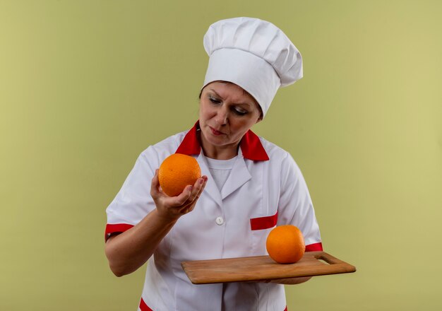 Thinking middle-aged female cook in chef uniform holding orange on cutting board and looking at orange in her hand 