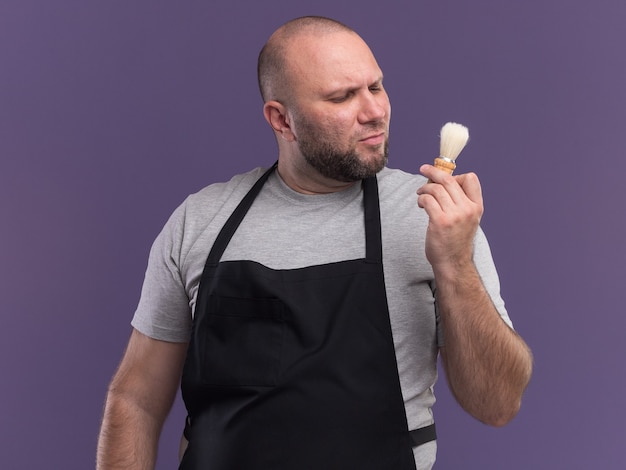 Thinking middle-aged male barber in uniform holding and looking at shaving brush isolated on purple wall