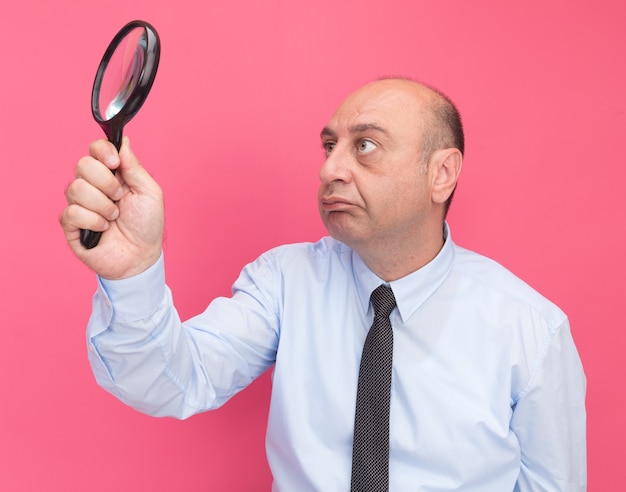 Free photo thinking middle-aged man wearing white t-shirt with tie raising and looking at magnifier isolated on pink wall