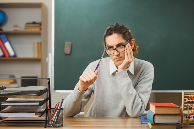 Free photo thinking putting hand on chin young male teacher wearing glasses holding pointer sitting at desk with school tools on in classroom