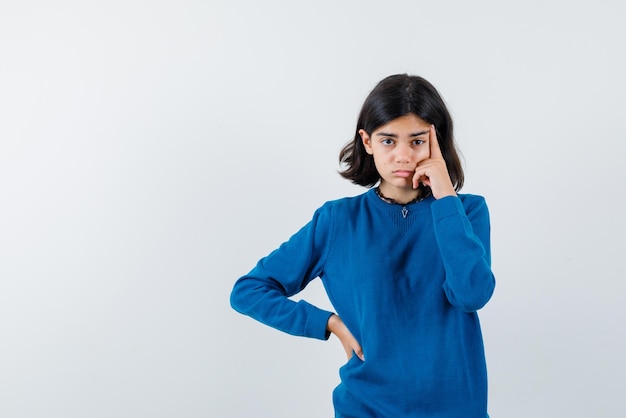 The thinking teenage girl is putting forefinger on temple and other hand on waist on white background