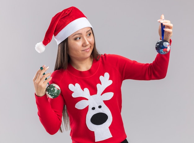 Free photo thinking young asian girl wearing christmas hat with sweater holding and looking at christmas tree balls isolated on white background