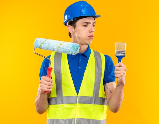 Thinking young builder man in uniform holding and looking at roller brush with paint brush 