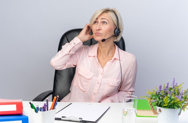 thinking young female call center operator wearing headset sitting at table with office tools isolated on white wall