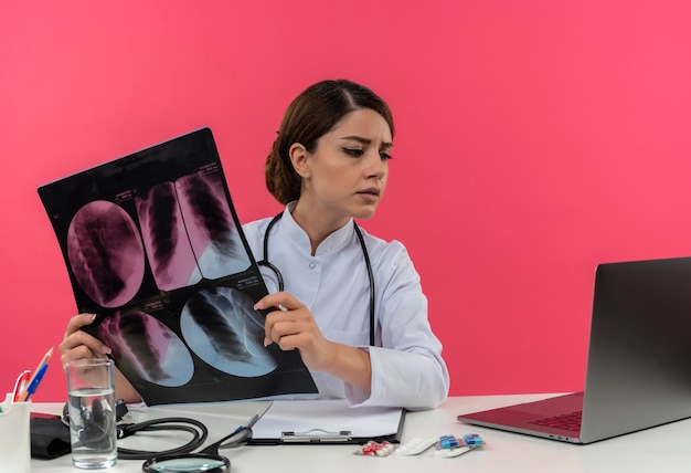 Thinking young female doctor wearing medical robe with stethoscope sitting at desk work on computer with medical tools holding x-ray and looking at laptop with copy space