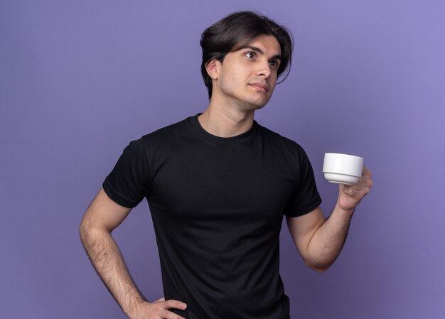 Thinking young handsome guy wearing black t-shirt holding cup of coffee putting hand on hip isolated on purple wall