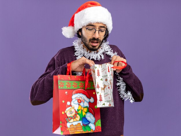 Thinking young handsome guy wearing christmas hat with garland on neck holding and looking at into gift bag isolated on blue background