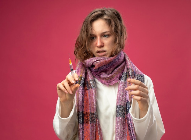 Free photo thinking young ill girl wearing white robe and scarf holding ampoule and looking at syringe in her hand isolated on pink wall