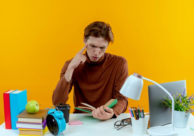 Thinking young student boy sitting at desk with school tools holding and looking at book putting hand on chin on yellow